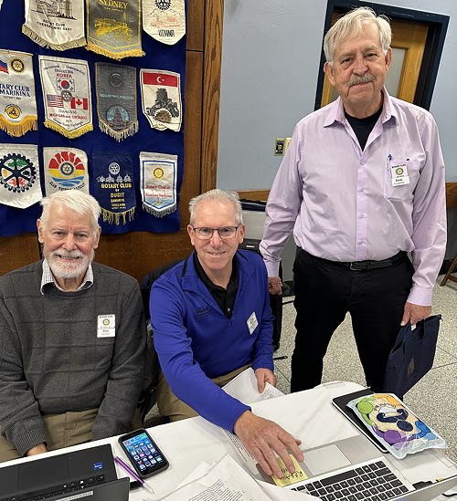 Three men, two sitting at a desk and one standing.