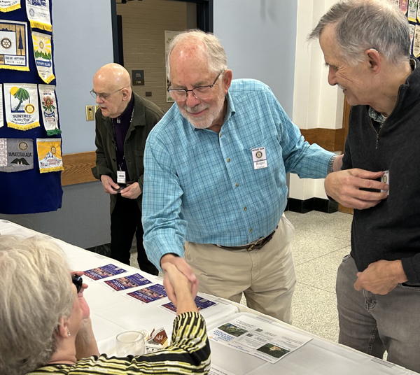 Men standing at table. One of them shaking hands with someone at the table.